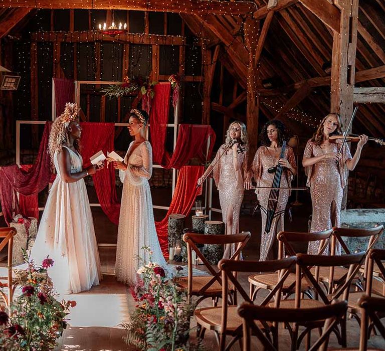 Brides reading their vows in front of square wooden decor with red and burnt orange wedding drapes, foliage and autumnal dried flower decor, fairy lights, festoon chandelier and string trio