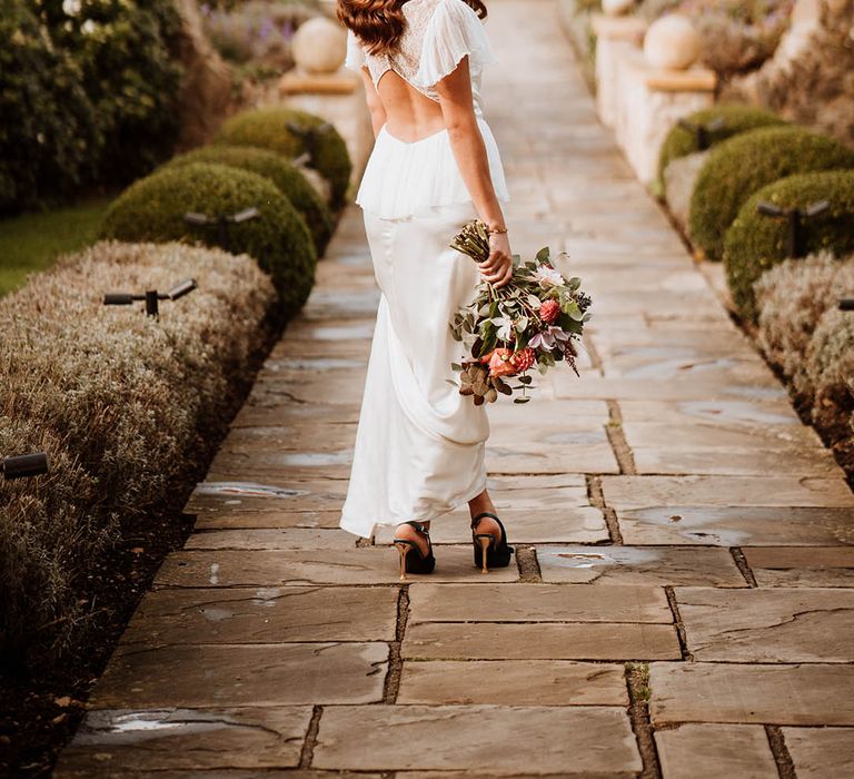 Bride holding wedding bouquet with white and pink carnations, garden roses, eucalyptus, baby’s-breath and dried flowers wearing satin open back wedding dress with button details and butterfly sleeves, ruffled and layer on the back and strappy black high heels