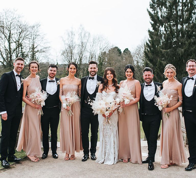 Wedding party shot of the bridesmaids in neutral pink dresses in different styles with the groomsmen and groom in black tuxedos and the bride in a fitted lace wedding dress 