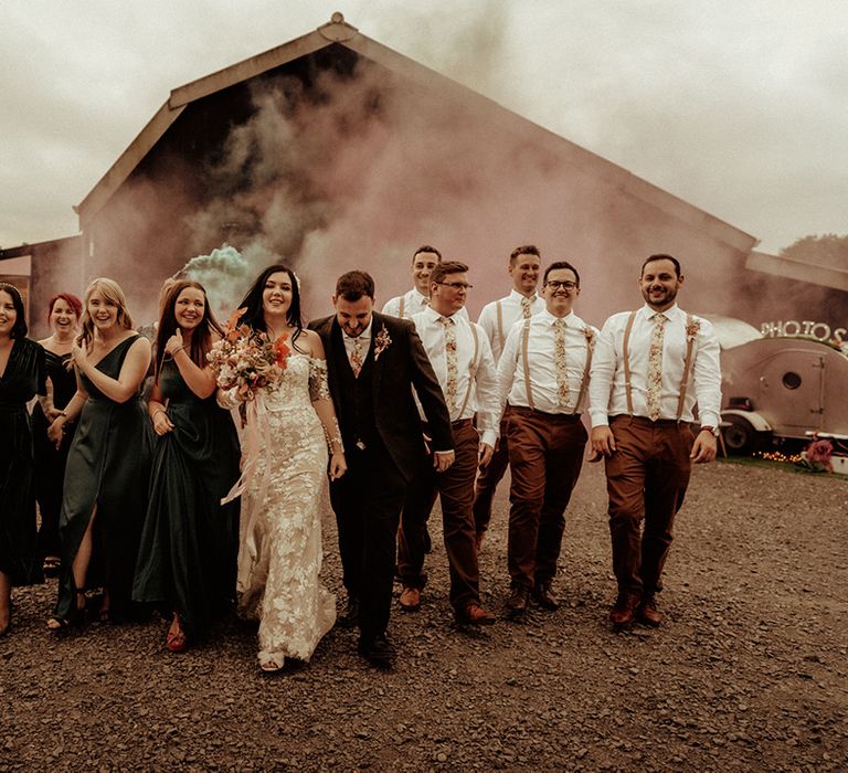 Bride in off the shoulder Abigail Westrup bridal gown with long, sheer sleeve and lace detailing, groom in dark suit, bridesmaids in dark green mismatched bridesmaid dresses and groomsmen in matching ties and suspenders doing wedding smoke bomb photo