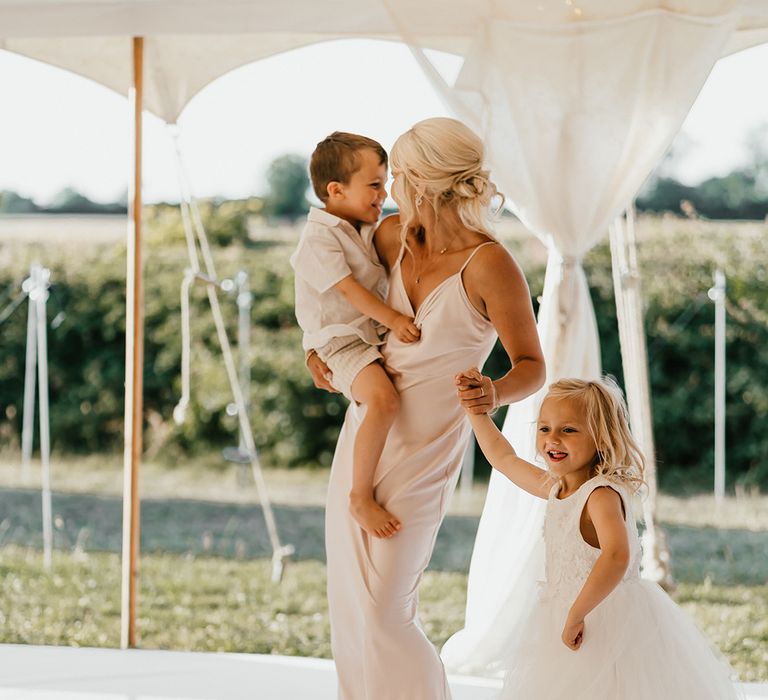 Blonde bridesmaid in white satin cowl neck dress dances with children during marquee wedding reception 