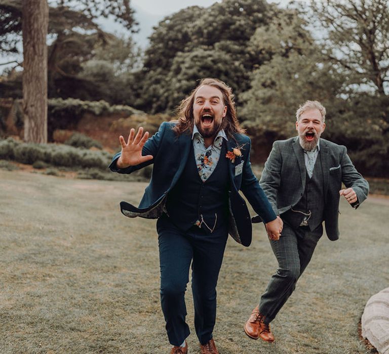 Groom in blue suit and floral shirt hold hands alongside his groom whilst running with one another outdoors 