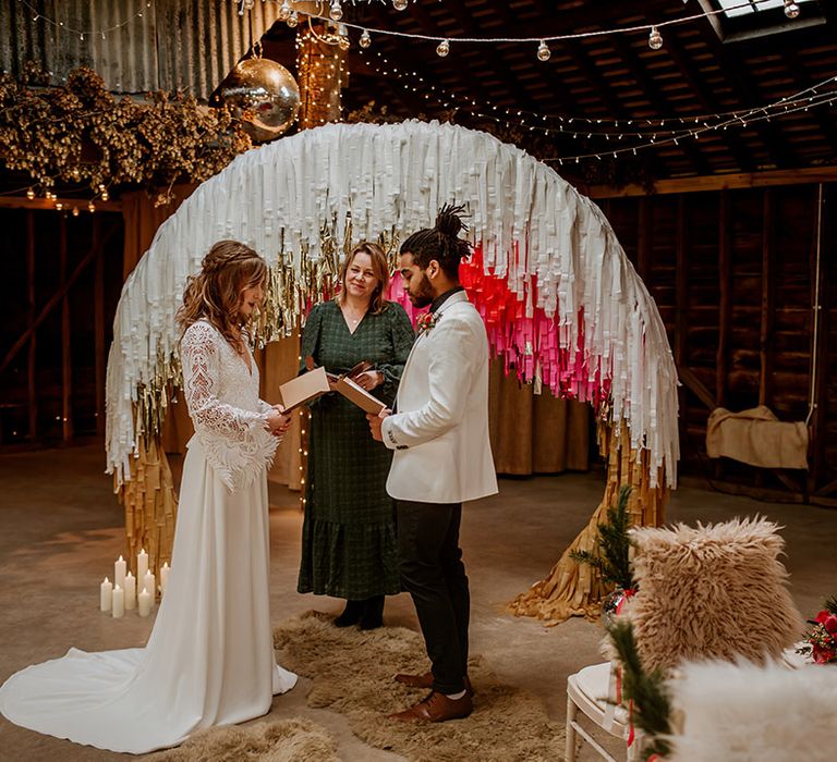 Groom in white blazer with colourful boutonniere, black shirt and black suit trousers and bride in boho long sleeve lace wedding dress with tassels reading vows to each other with retro decor around them; disco ball decor, fairy lights, dried flowers and large semi-circle arch decor with white, gold and hot pink streamers 