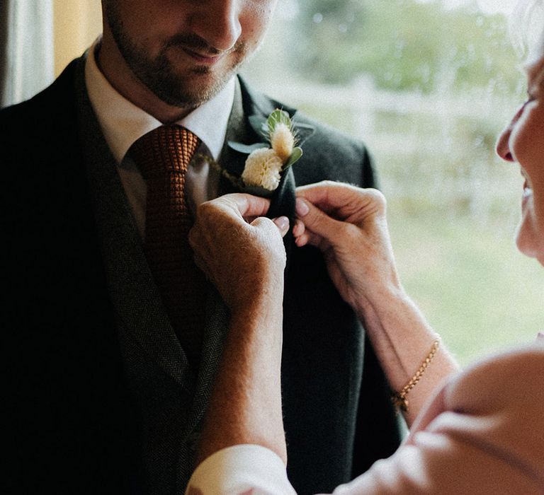 Groom wearing his wedding suit with an orange tie stands as the mother of the groom attaches the buttonhole 