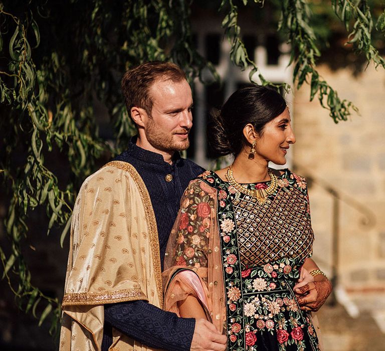 Bride wears beautifully embellished lehenga as her groom stands beside her wearing golden sash across velvet jacket during golden hour portraits