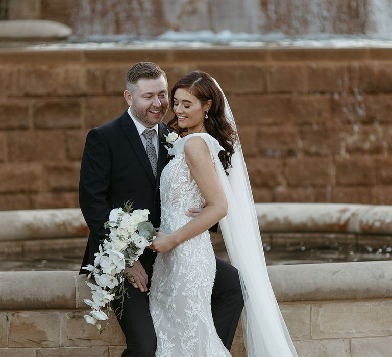 Groom in black suit sits on the edge of the fountain with the bride in a fitted floral wedding dress holding an all white cascading bouquet 