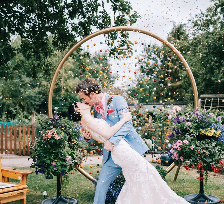 Bride in lace wedding dress kissing the groom in a light blue suit with pom pom moongate decoration for outdoor wedding 