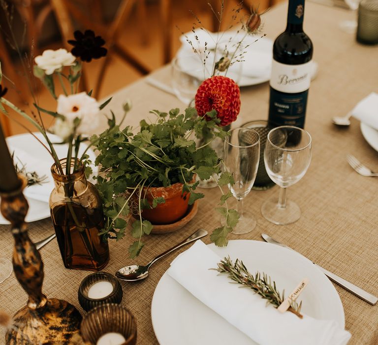 Red dahlia and potted plant with flowers in a vase and small tea lights with white plates decorated with sprig of rosemary place names and napkin