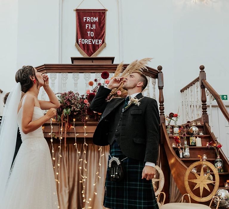 Bride in Stephanie Allin bridal gown and groom in a kilt drinking whiskey shots at their bespoke wedding ceremony 