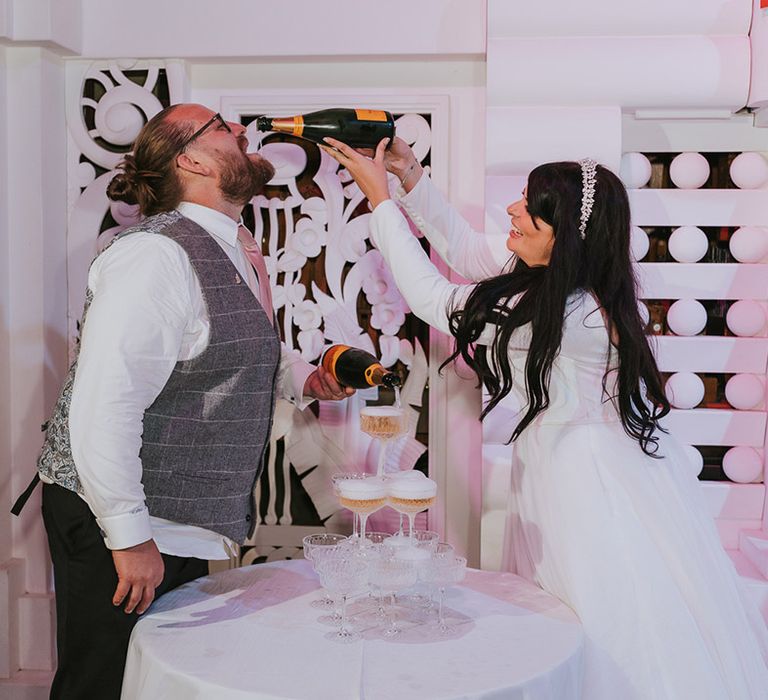 The bride pours champagne into the groom's mouth as he pours champagne onto the champagne tower decoration 