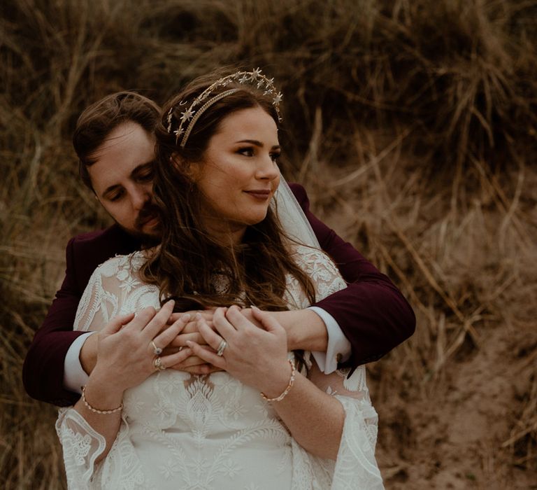 Bride in a boho lace bell sleeve wedding dress from Rue De Seine with a gold bridal crown being embraced from behind by the groom in a burgundy suit 