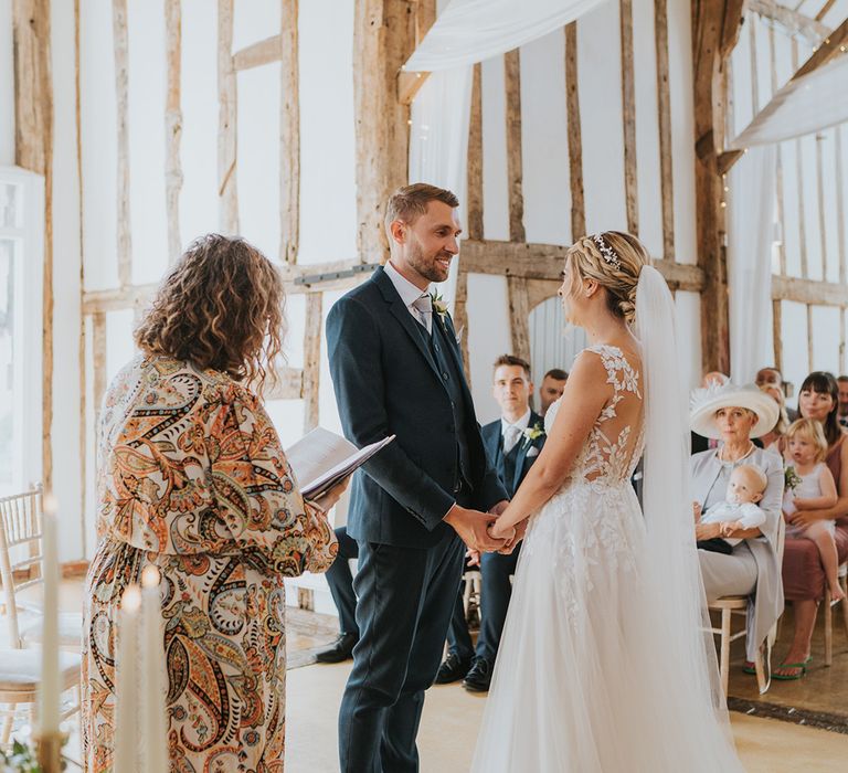 Groom in three piece navy suit stars facing the bride holding her hands in an open back illusion wedding dress for rustic barn wedding 