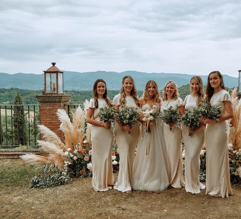 Bridal party pose at altar wearing off-white satin bridesmaid dresses in front of pampas grass decor whilst holding eucalyptus bouquets