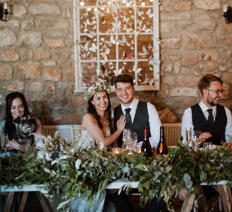 Bride in white garden rose bridal flower crown sitting with groom in dark grey waistcoat and tartan tie at The Barn at Harburn wedding venue with foliage centrepieces and 3d paper butterfly decorations