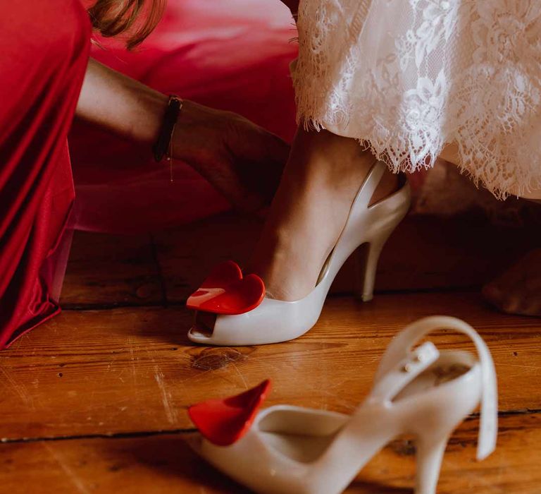 Bridesmaid in red bridesmaid dress helping bride into white Vivienne Westwood wedding heels with red heart on top 