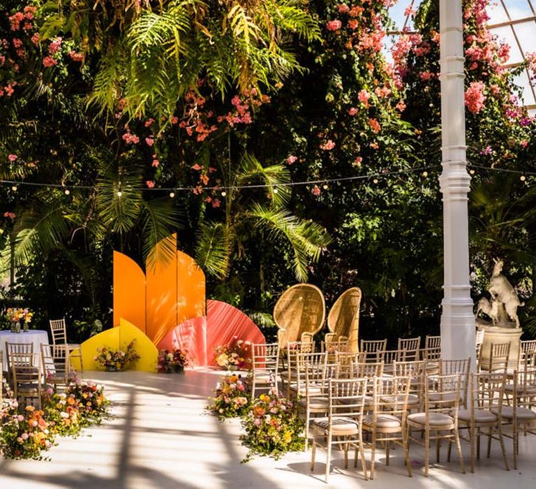 Botanical wedding venue decorated with chairs, colourful aisle wedding flowers, and orange, yellow and pink arched altar decor 