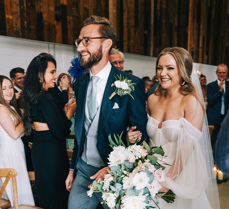 Groom wearing a white flower buttonhole in navy suit walking with the bride in off the shoulder long sleeve Alena Leena wedding dress 