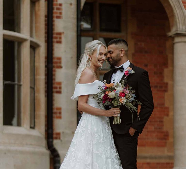 Groom in classic three piece grooms tuxedo with colourful spring boutonniere standing with bride in off the shoulder lace wedding dress holding colourful spring bridal bouquet outside Frensham Hall Manor House wedding venue 