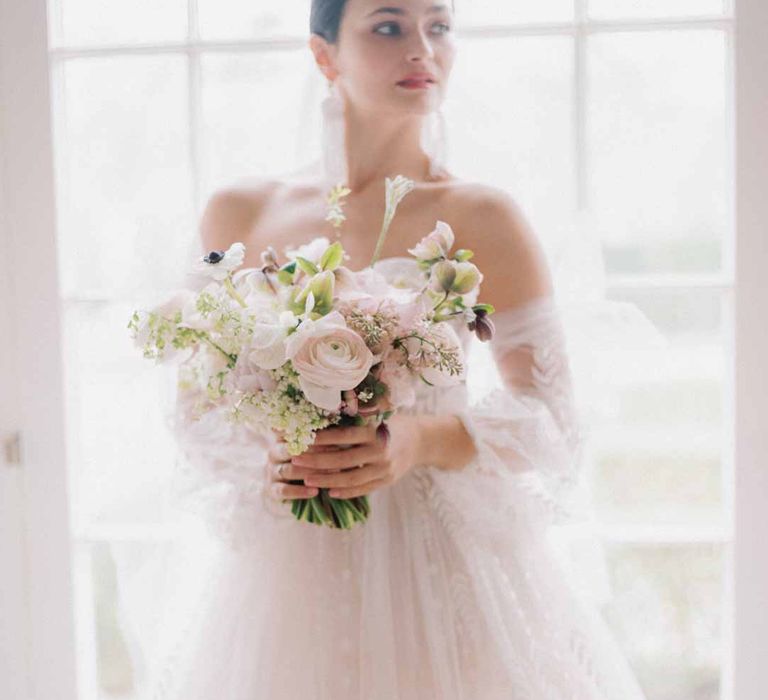 Bride standing by window at Settrington Orangery in sweetheart neckline tulle mesh bridal gown with detachable puff tulle sleeves and delicate beading holding bridal bouquet with white poppies, light pink Persian buttercups, baby's-breath and foliage 