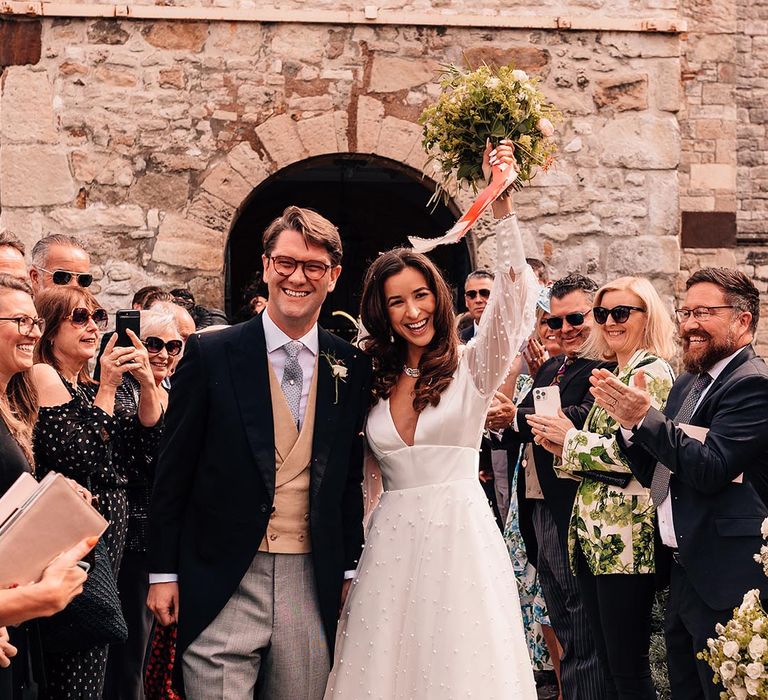 Bride in pearl wedding dress with sheer sleeves with the groom in a morning suit with blue tie and yellow waistcoat 