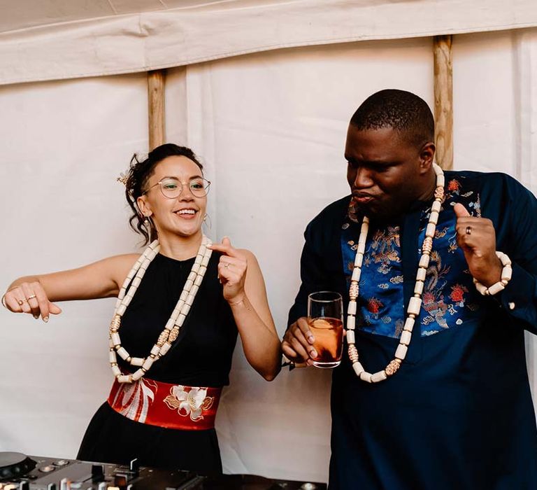 Bride in black bridal jumpsuit with red and gold Chinese fabric belt dancing with groom in traditional Nigerian wedding outfit both wearing white beads around their neck at Broadfield Court