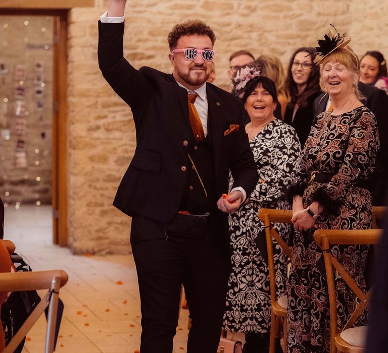 Wedding guest wearing dark suit with burnt orange wedding tie and pocket square walking down the aisle at Kingscote Barn Tetbury 