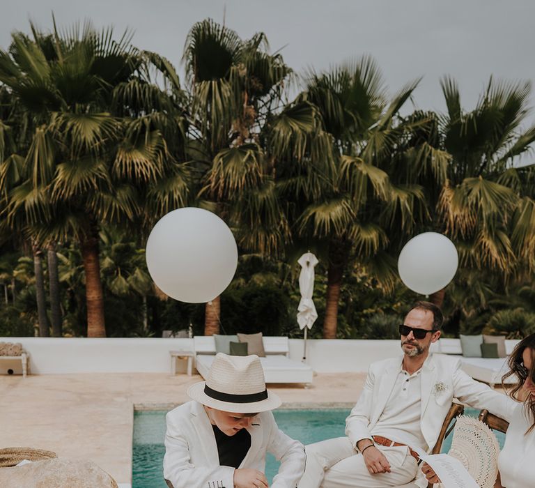 little boy ring bearer in a white suit and trilby hat at poolside ibiza wedding ceremony