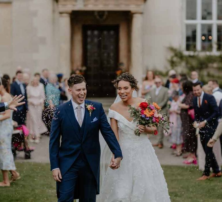 Bride in Suzanne Neville off shoulder 3D floral applique wedding dress walking hand in hand with groom in navy groom morning suit with grey ties and blush rose boutonniere doing colourful wedding confetti exit