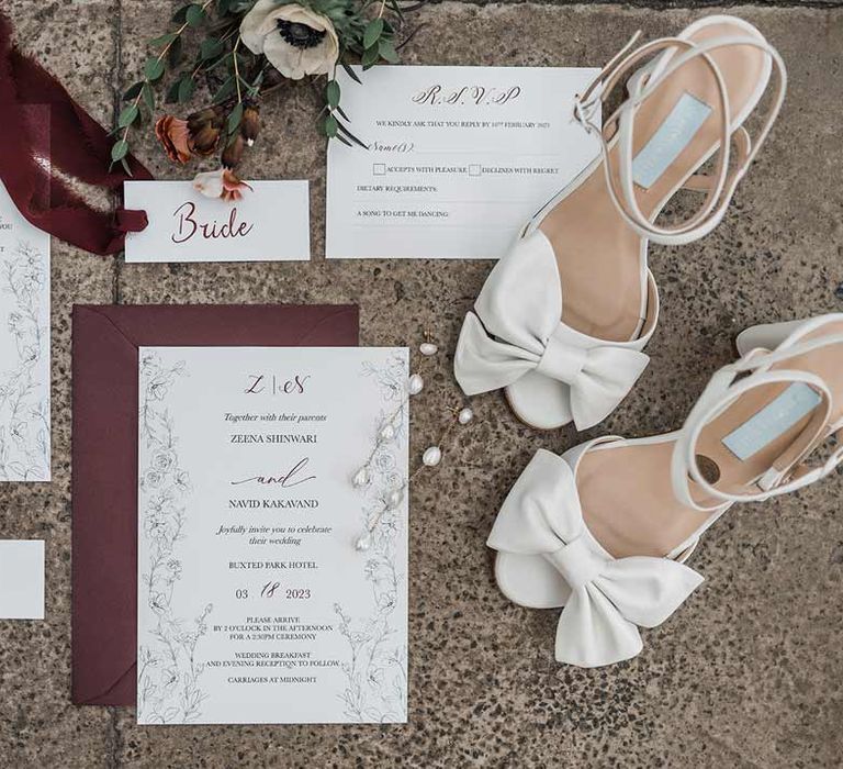 Minimalist white and maroon wedding stationery with fine line drawings and calligraphy surrounded by dried flowers, maroon ribbon and white open-toed wedding heels