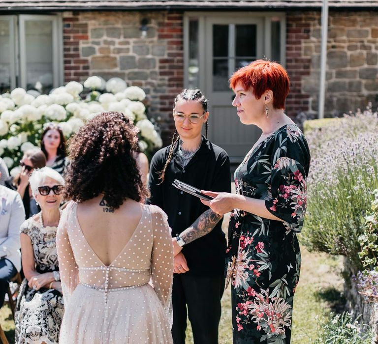 Celebrant leading handfasting ceremony at Viking themed wedding for bride in black shirt and black trousers and bride in long sleeve v-neck sheer polka dot overlay sparkly wedding dress and gold celestial bridal headband 