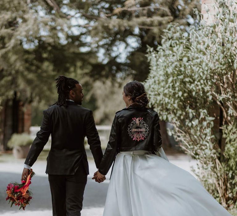 Bride in strapless sweetheart neckline wedding dress and personalised bridal leather jacket holding hands with groom in classic black tuxedo 