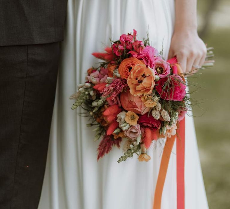 Bride holding colourful pink and orange wedding bouquet with poppies, garden roses, carnations, foliage, and dried flowers 