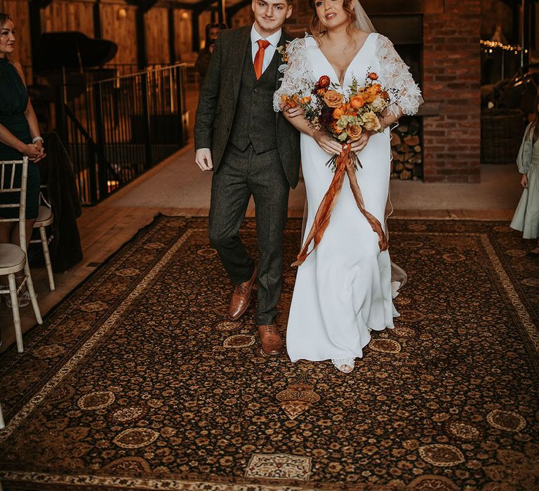 Bride in boho wedding dress being walked down the aisle with her son holding an autumnal bouquet 