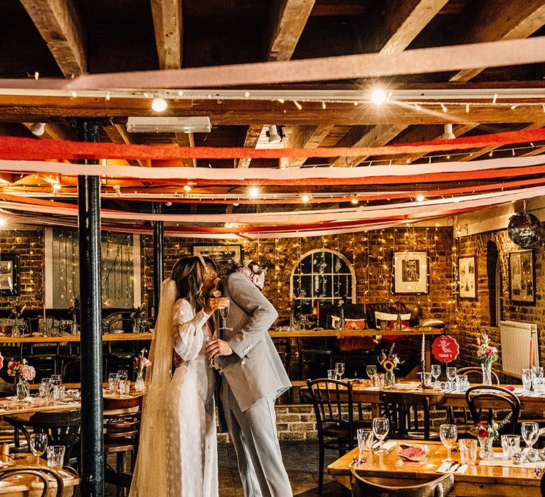 bride in a jumpsuit and groom in a grey suit embracing in the middle of their wedding reception with streamer ceiling decor at East Quay wedding venue 