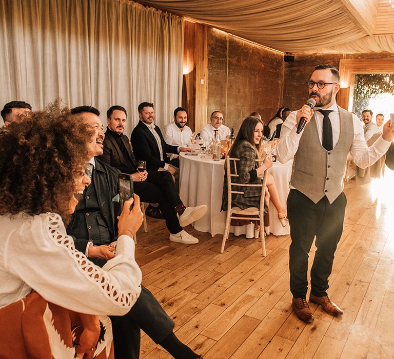 The groom stands up to deliver a wedding speech with guests laughing and filming on their phones 