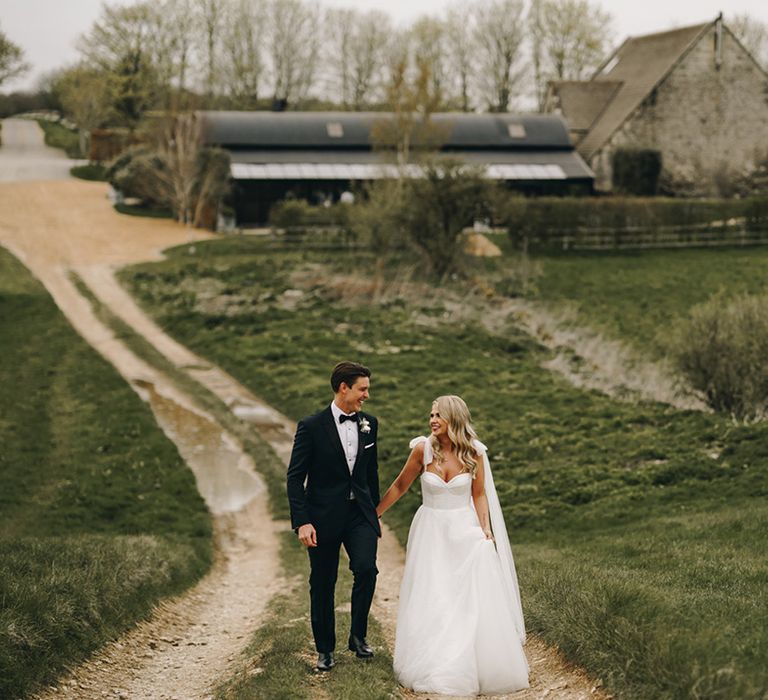 Bride in sparkly wedding dress in bow tied straps walking with the groom in a black tuxedo around the Stone Barn venue in the Cotswolds