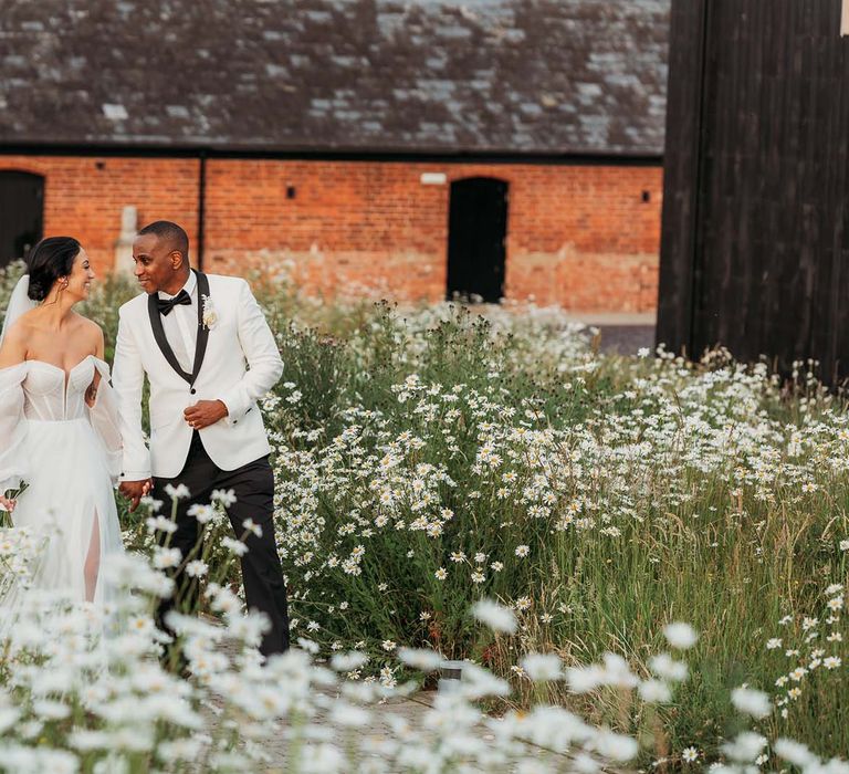 Bride in off-the-shoulder wedding dress walks through daisy gardenw ith groom in black and white tuxedo at black-tie wedding