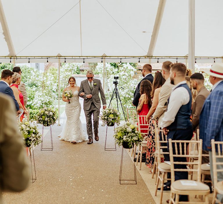 The father of the bride in a grey suit starts to walk down the aisle with the bride at marquee ceremony 