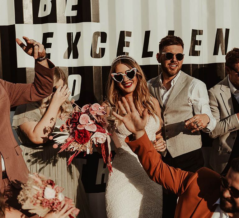 Bride in lace gown carrying a pink bouquet with the groom wearing rounded black sunglasses 