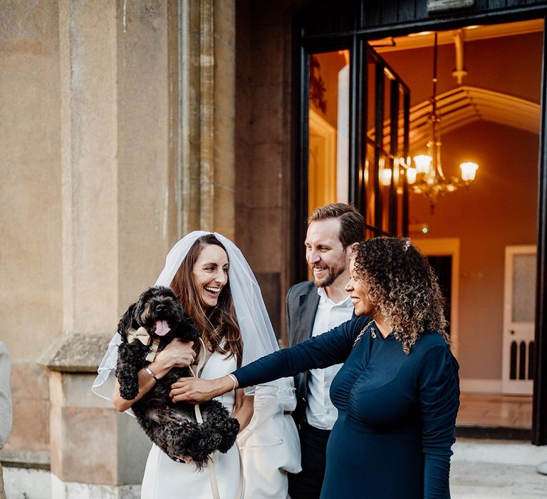 Bride walks with wedding guests carrying a pet dog wearing a bow collar 