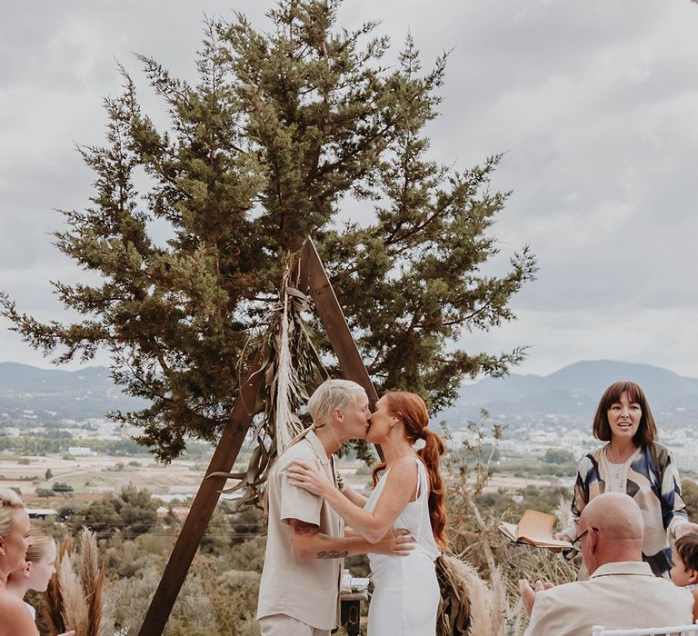 Two brides share their first kiss together as they're announced as a married couple at their same sex outdoor wedding ceremony 