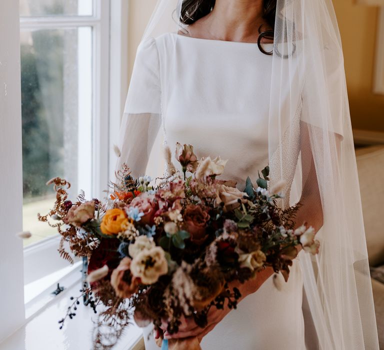 Bride laughs to camera. She wears an embellished dress and veil and holds a winter bouquet. 