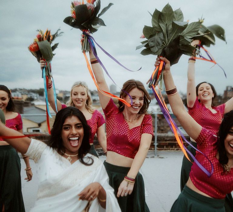 Bride in a white sari and bridesmaids in pink and green Lenghas holding their colourful bouquets in the air 