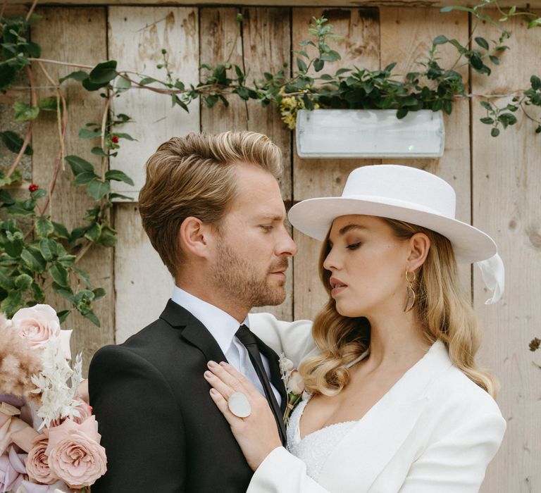 Groom in a black suit embracing his bride in a sequin jumpsuit, blazer and bridal hat