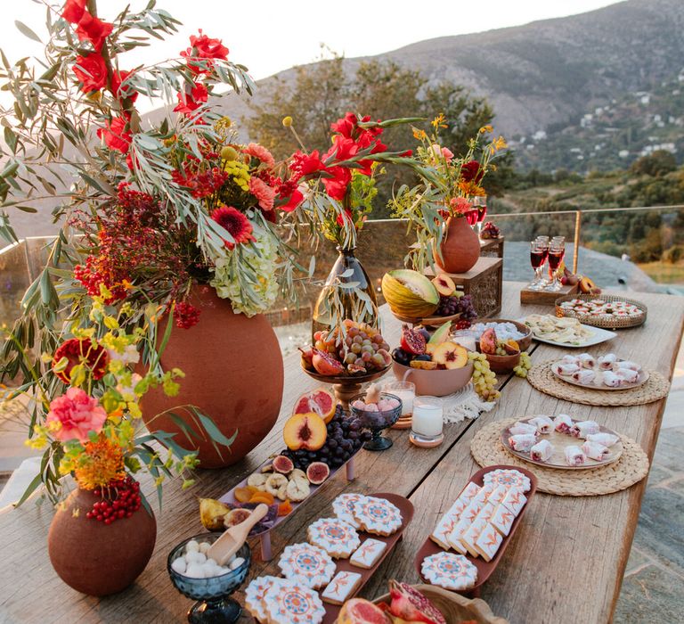 A grazing table full of local Greece produce including cookies, fruit and desserts, and terracotta vases with wildflowers