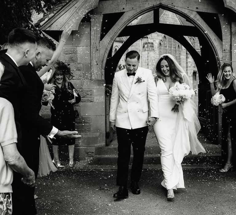 Smiling groom in white tuxedo jacket walks through confetti with smiling bride in white Made With Love wedding dress and white chapel length veil