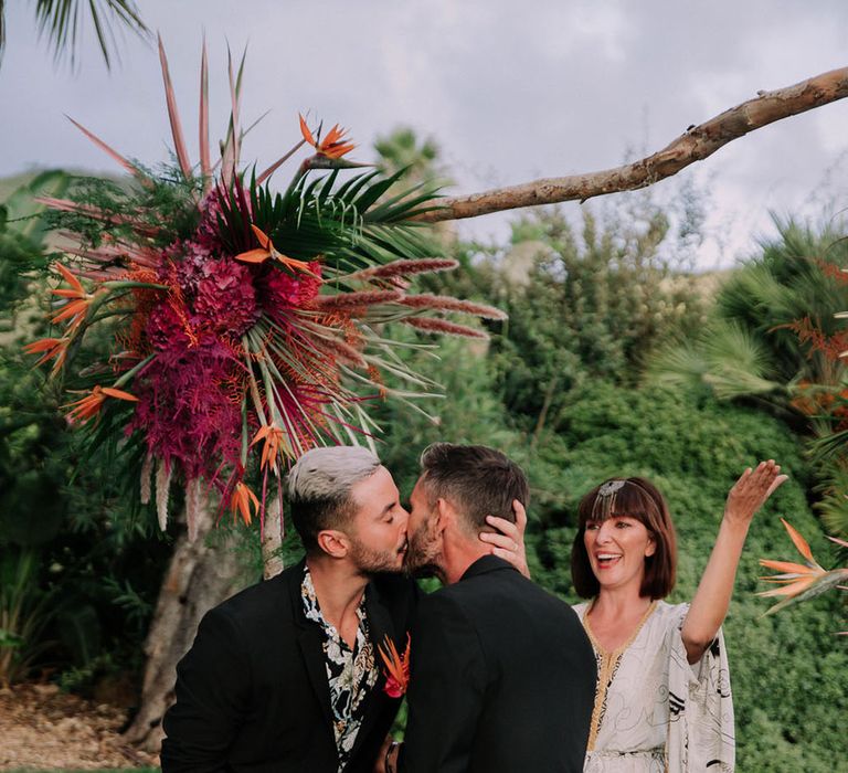 Grooms kiss under a wooden altar at drag race inspired wedding. Photography by Stephanie Shenton.