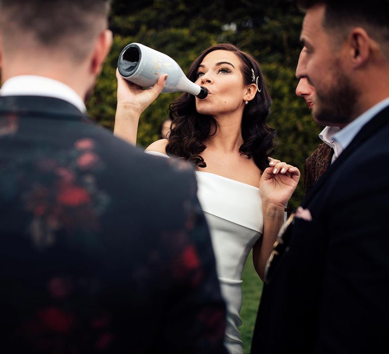 Bride in white strapless Vagabond wedding dress wearing celestial hair pins swigs from white bottle at lakeside wedding