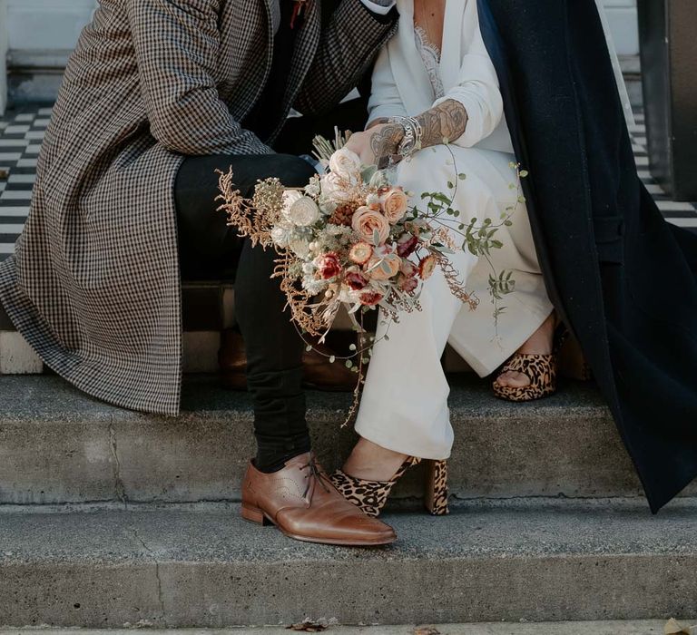 White bride in white suit with green coat over shoulders kisses groom with tweed jacket