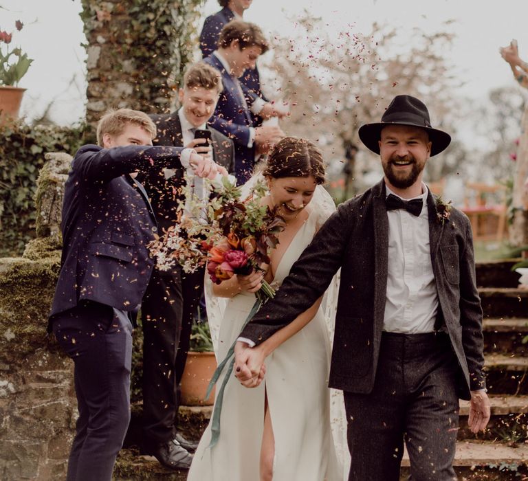 Laughing groom in grey tweed suit and black fedora holds hands with laughing bride in Charlie Brear wedding dress and applique veil walking down garden steps whilst guests throw confetti at garden party wedding in Devon